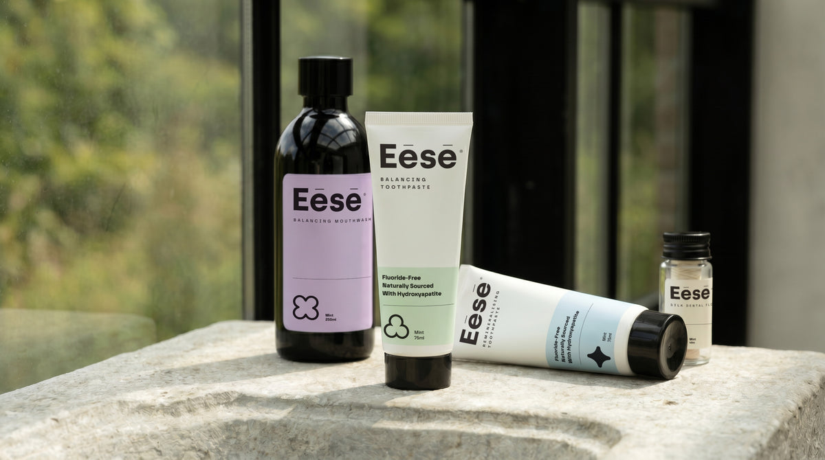 Eēsē oral care set featuring sodium lauryl sulfate free toothpaste tubes, a sleek black mouthwash bottle, and natural dental floss on a stone surface in front of a window.