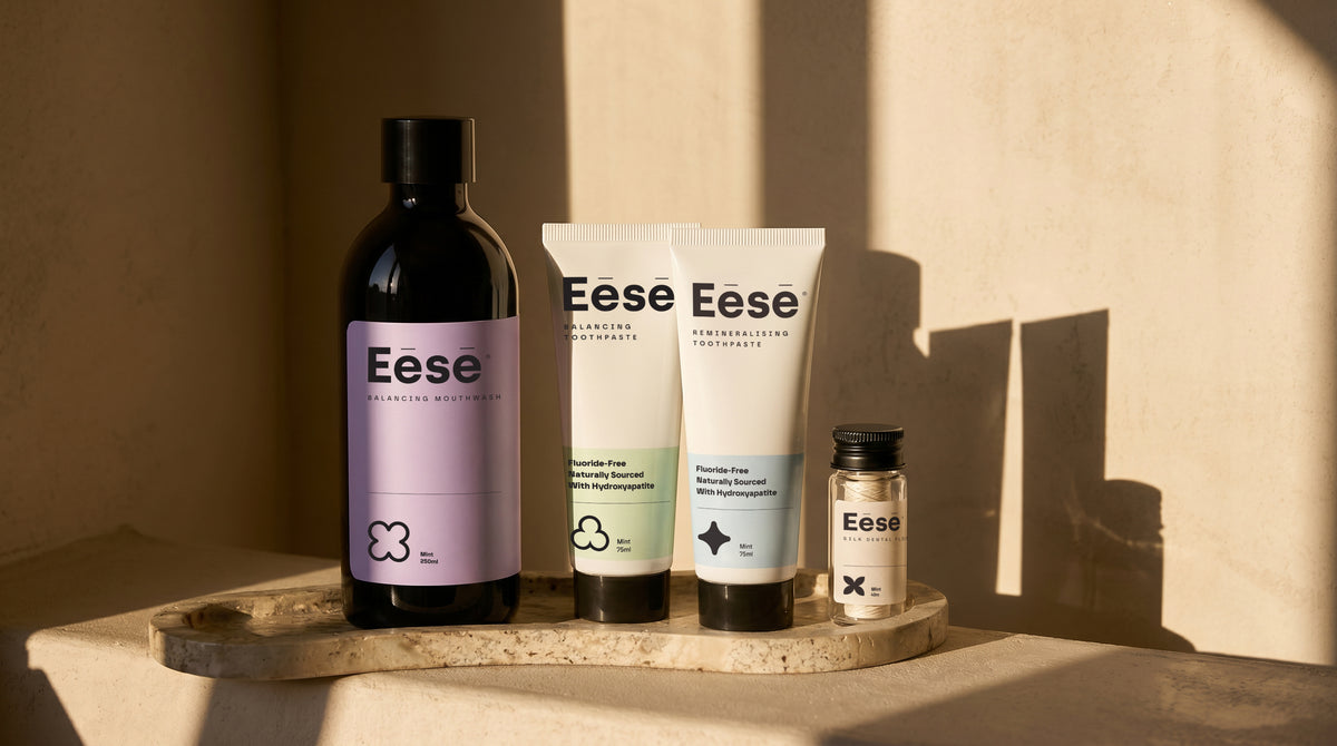 Eēsē complete oral care collection — natural toothpaste without SLS, alcohol-free mouthwash, and the best floss for daily use, arranged on a on a stone tray in warm, direct sunlight against a beige wall.