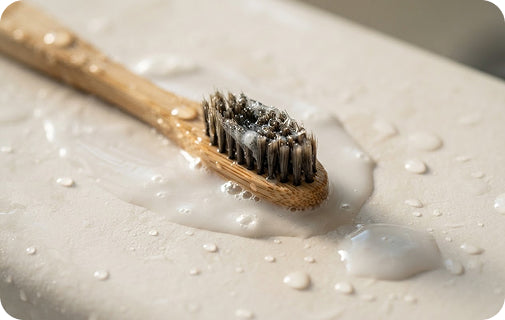 A close-up of a light natural wood toothbrush with dark bristles, resting on a white textured surface covered in water droplets and a small pool of white foam from all natural toothpaste.